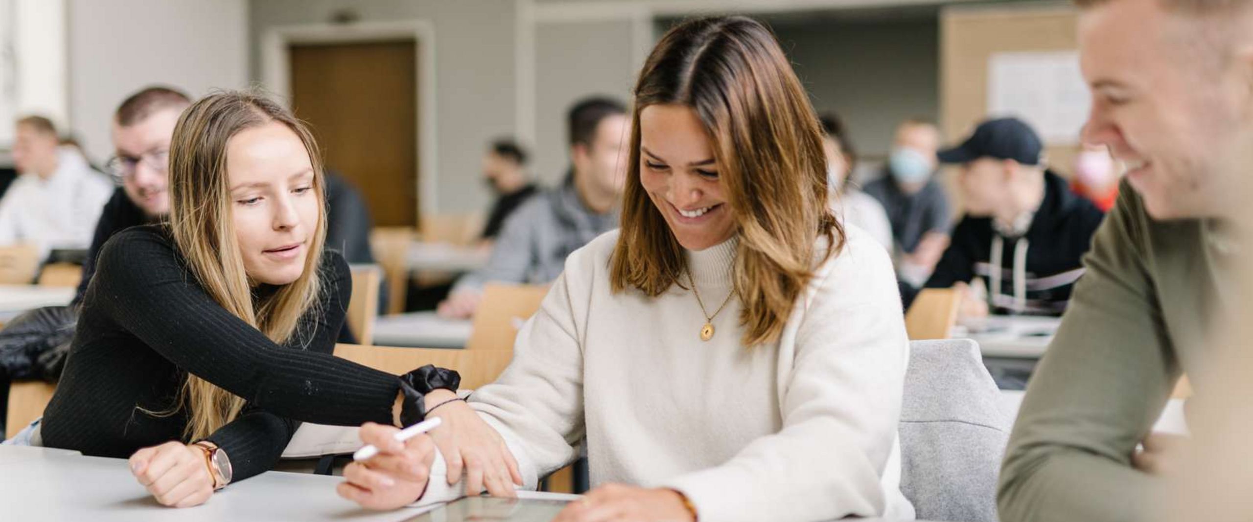 Studierende im Seminar Zwei Studentinnen und ein Student sitzen an einem Tisch in einem Seminarraum. Im Hintergrund sieht man viele weitere Studierende. Die Studentin in der Mitte hat ein Tablet vor sich, einen Tablet-Stift in der Hand und lacht. Die Studentin neben ihr zeigt auf etwas auf dem Tablet. Der Student neben ihr schaut den beiden zu und lacht.