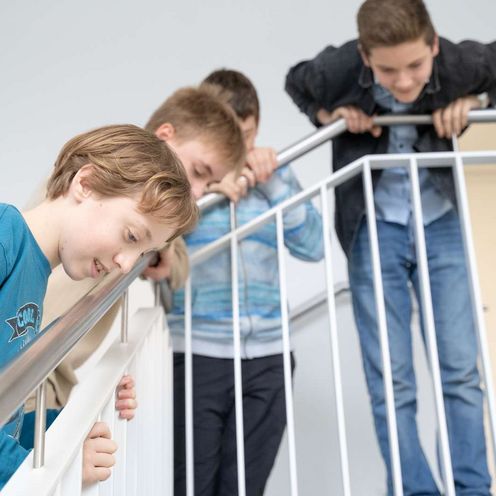 Beobachten Auf dem Bild ist eine Gruppe von Schüler:innen zu sehen, die sich gespannt über das Treppengeländer nach unten beugen, um unten etwas zu beobachten.