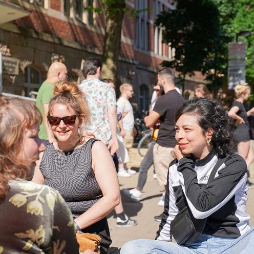 Studentinnen vorm M-Gebäude am Campus Neustadt Drei Studentinnen sitzen bei sonnigem Wetter auf einer Bank vorm M-Gebäude am Campus Neustadt und reden miteinander. Im Hintergrund steht eine Gruppe Studierender und unterhält sich ebenso.