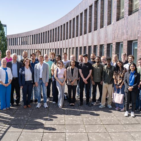 Teilnehmende der EMSS Industry Week 2025 Auf dem Foto sind rund 30 Teilnehmende zusehen. Sie stehen in einer Gruppe zusammen auf einer Terrasse. Im Hintergrund ist ein dunkelrotes Backsteingebäude zu erkennen. Die Sonne scheint.