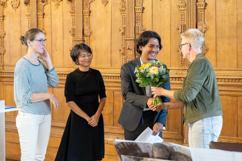 Frau schüttelt jungem Mann die Hand, der einen Blumenstrauß hält. Zwei Frauen schauen lächelnd zu.