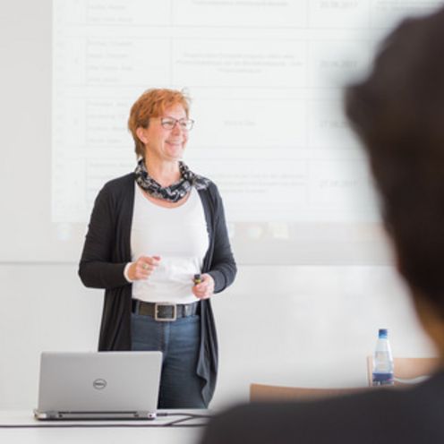 Dozentin Eine Dozentin steht vor einer Wand, auf die eine Präsentation projiziert wird. Sie spricht zu Studierenden in einem Seminarraum.
