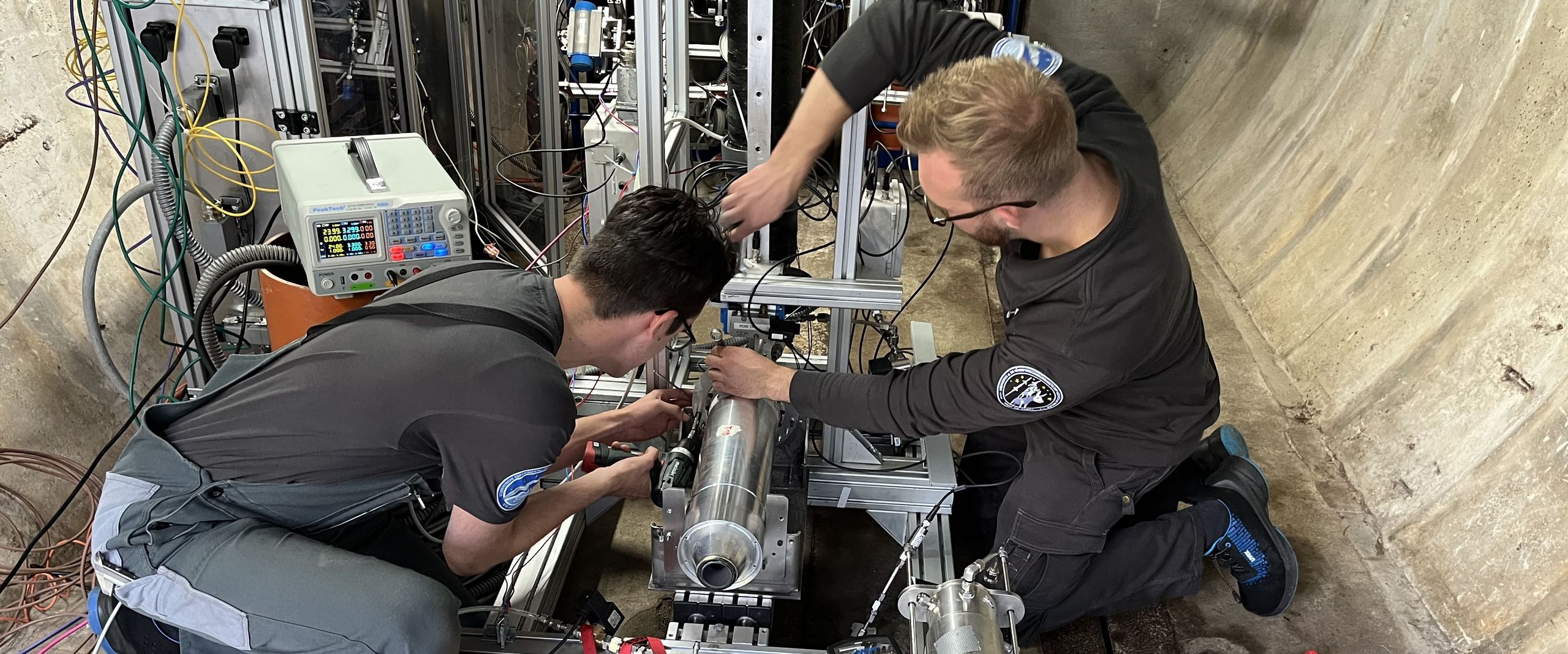 Auf dem Bild sind die Vorbereitungen des Triebwerktests zusehen. Zwei Studenten sitzen vor dem Triebwerk in einer großen Betonrühre und schrauben an dem Triebwerk herum. 