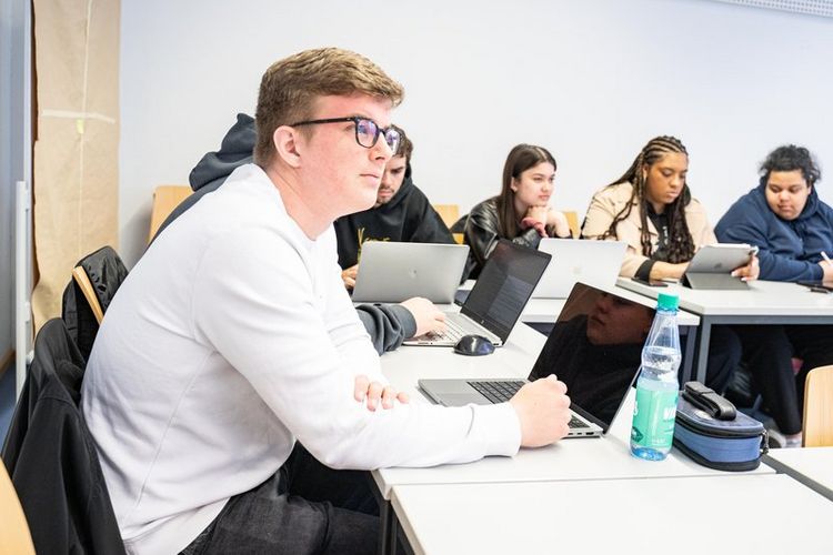 Studierende im Seminar Studierende sitzen über Eck an Tischen in einem Seminarraum. Sie schauen teils nach vorn, teils auf ihre Notizen oder Notebooks, die vor ihnen platziert sind.