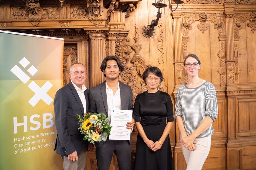 The photo shows four people looking into the camera. One is holding flowers and a certificate