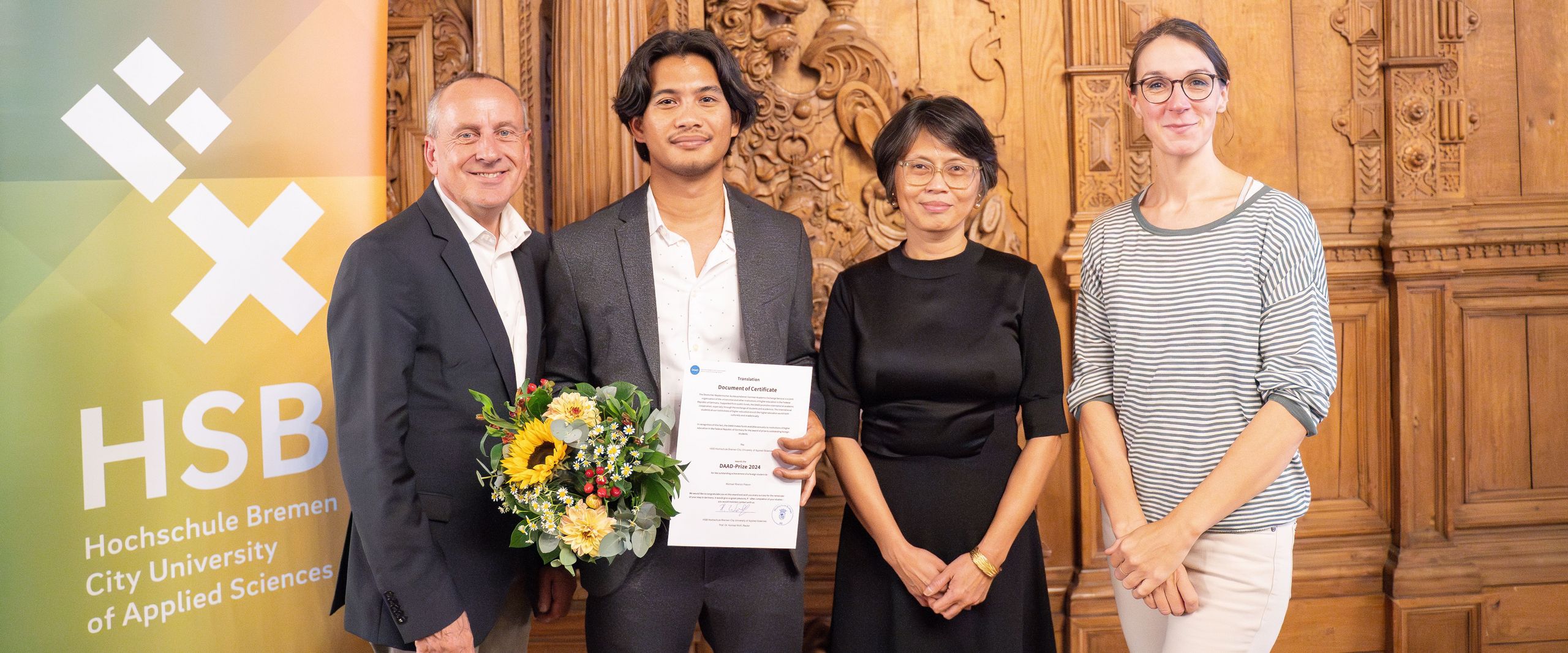 The photo shows four people looking into the camera. One is holding flowers and a certificate