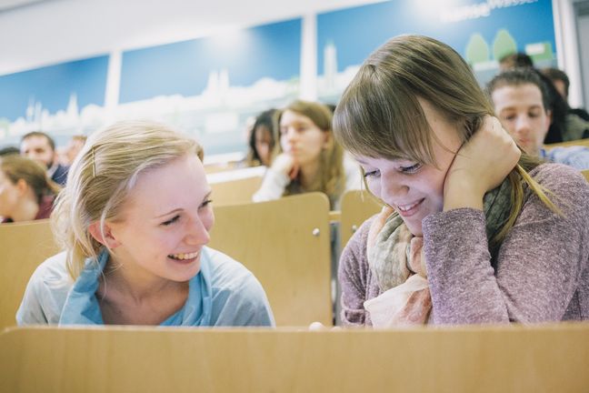 RS432__MG_5909.jpg Zwei junge Frauen sitzen einander zugewandt und lächelnd im Hörsaal.