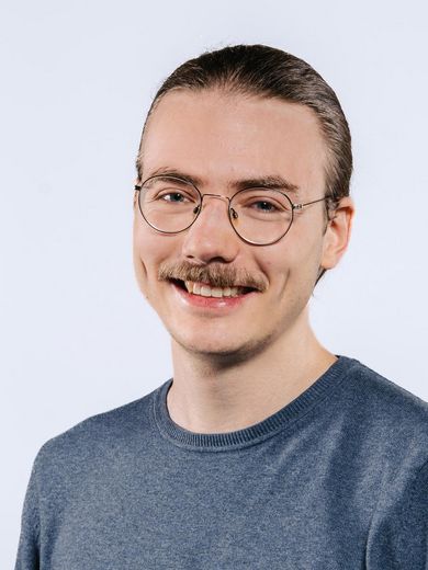 Daniel Hackbarth Auf dem Foto ist Daniel Hackbarth zusehen. Er hat seine braunen Haare zu einem Zopf zusammengebunden. Er trägt eine runde Brille und einen blauen Pullover.