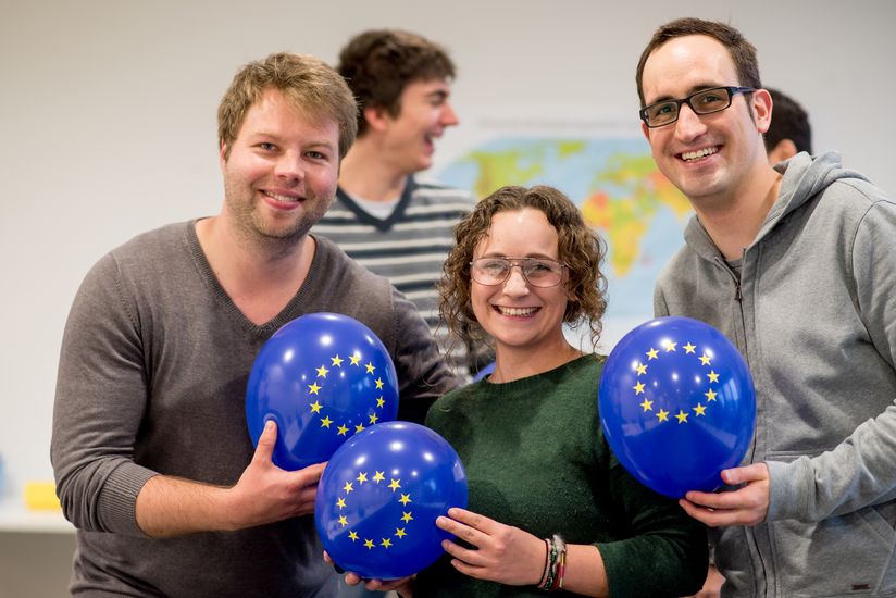 Zwei junge Männer und eine junge Frau halten dunkelblaue Luftballons mit den EU-Sternen und lachen in die Kamera.
