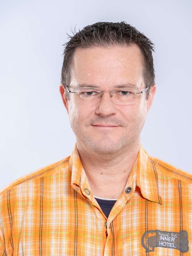 Auf dem Bild ist Andreas Albrecht zusehen. Er hat kurze schwarze hochgelegte Haare und trägt eine unauffällige Rahmenlose Brille und ein orange-kariertes Hemd