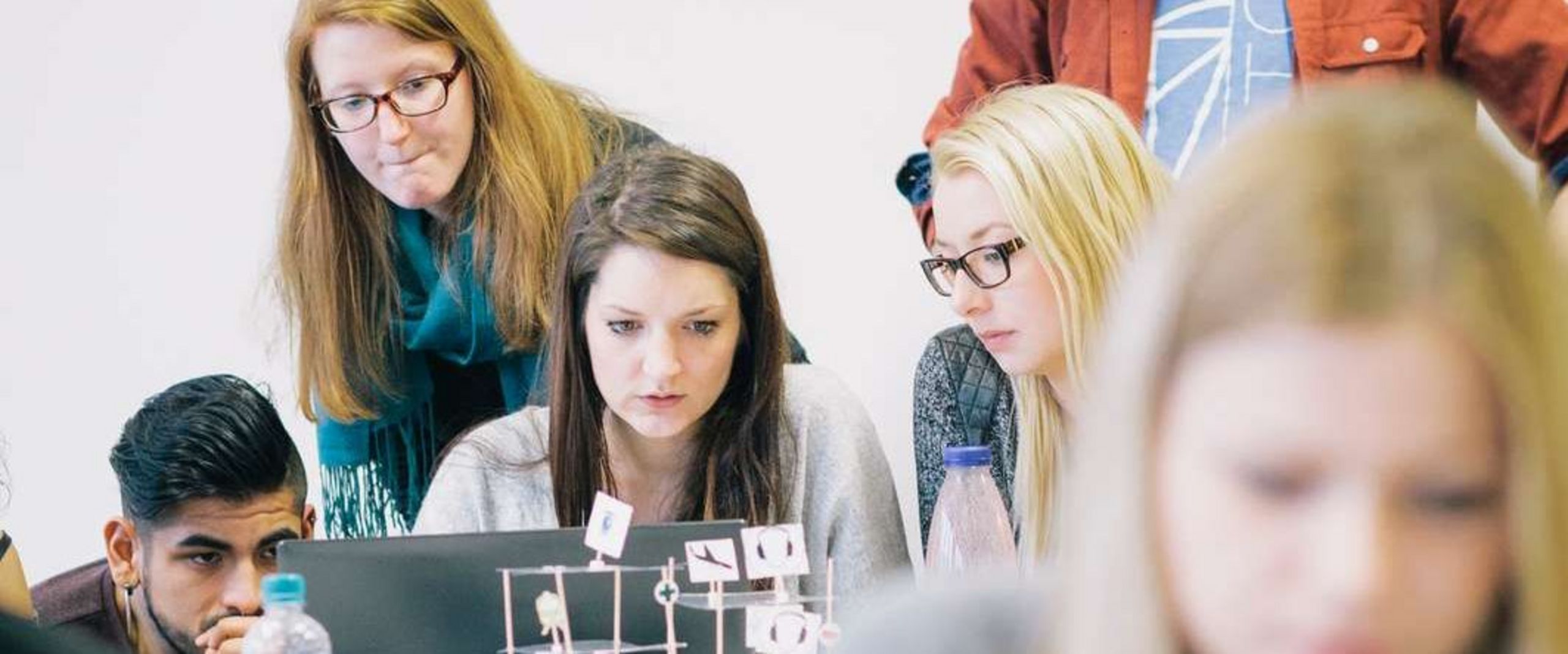Auf dem Bild ist eine Gruppe Studierender zu sehen, die sich rund um einen Laptop positionieren und nachdenklich aussehen. Im Vordergrund sitzt eine Studentin vor einem roten Laptop. Der Vordergrund ist unscharf. 