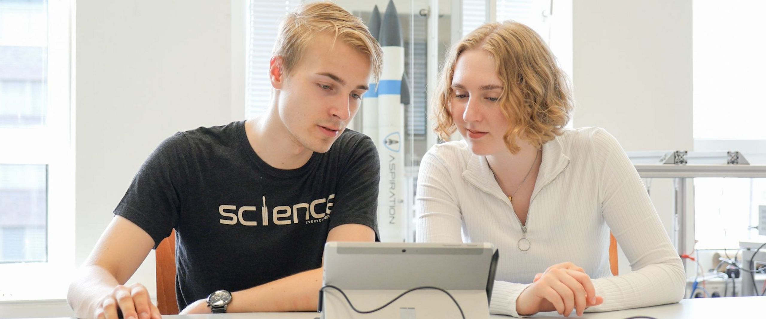 Auf dem Bild sind ein blonder junger Mann und eine blonde junge Frau zusehen, die vor einem Tablet sitzen. Hinter ihnen ist eine Rakete in einem Schaukasten. 