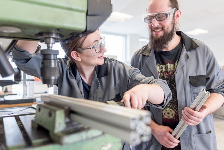 Werkstatt am ZIMT Eine Studentin spannt eine Metallstange in eine Maschine ein, während ein zweiter Student ihr dabei zusieht.