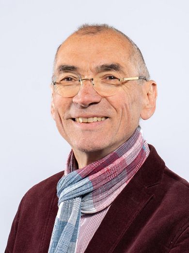 Peter Schmidt Auf dem Foto ist Peter Schmidt zusehen. Er hat sehr kurzes graues Haar. er trägt eine Brille, Ein dunkelrotes Jackett und einen rot-blauen Schal.