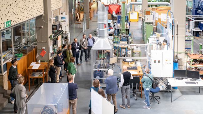 Rundgang in der WKL Halle Auf dem Bild ist eine Gruppe Leute fotografiert, die sich um einen Elektrolyseur versammelt haben. Das Bild ist aus der Vogelperspektive aufgenommen.