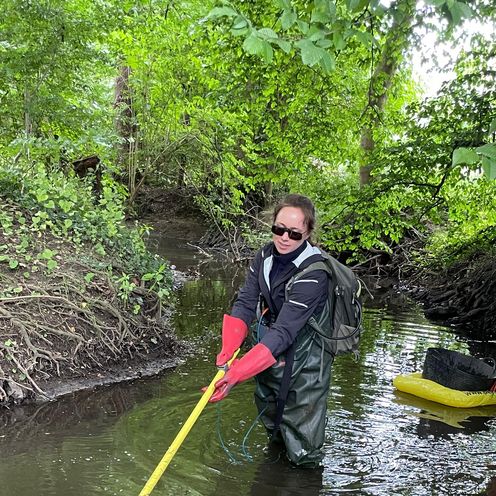 Isabel Tanzberger bei den Gewässeruntersuchungen Eine Frau mit Rucksack steht im wasserdichten Anzug in einem kleinen Wasserlauf. Das Wasser reicht bis zu den Oberschenkeln. Sie taucht eine gelbe Stange ins Wasser, eventuell ein Fischköcher. Im Hintergrund schwimmt ihre weitere Ausrüstung auf einer Luftmatratze.