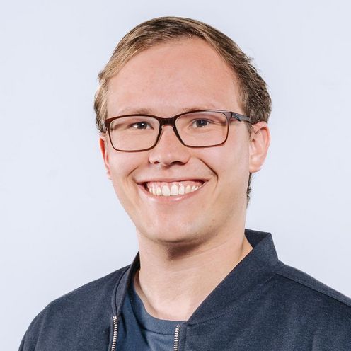 Tim Gust Auf dem Bild ist Tim Gust zusehen. Er hat blonde Haare und trägt eine Brille. Er hat ein blaues T-Shirt unter einer dunkelblauen Jacke an.
