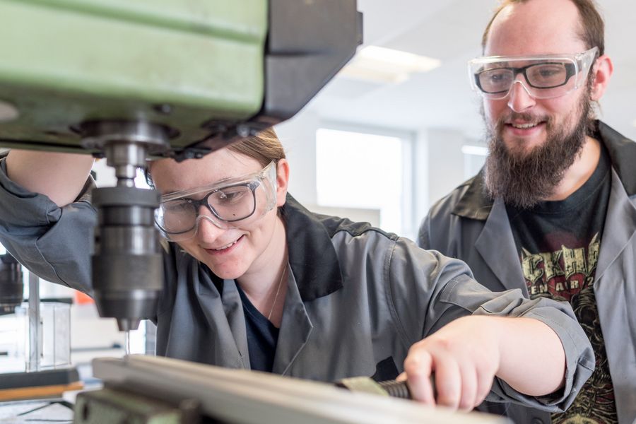 Werkstatt am ZIMT Eine Studentin spannt eine Metallstange in eine Maschine ein, während ein zweiter Student ihr dabei zusieht.