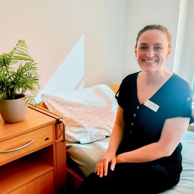 202508_TCALL Inia Tinis Frau sitzt auf einem Bett und lacht in die Kamera.