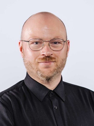 Auf dem Bild ist Nikolai Sokolowsky zusehen. Er hat eine Glatze und einen blond-rötlichen Bart. Er trägt eine Brille und ein schwarzes Hemd. 