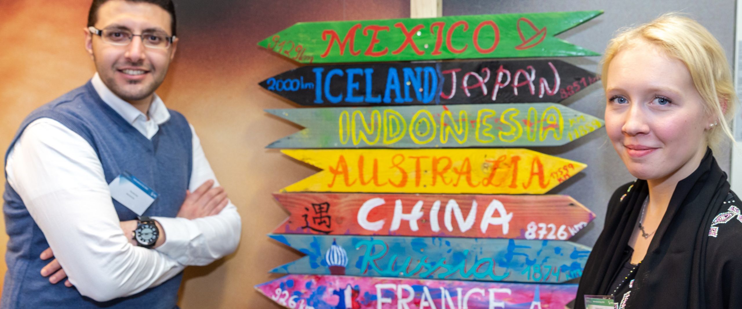 International Day 2018 Zwei Studierende posieren neben einem Wegweiser, auf dem verschiedene Länder stehen.