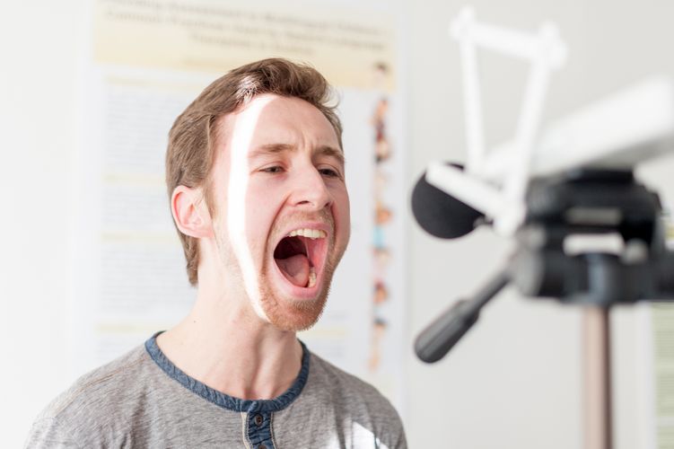 Übung Logopädie Ein Student spricht mit weit geöffnetem Mund in ein Mikrofon während einer logopädischen Übung.