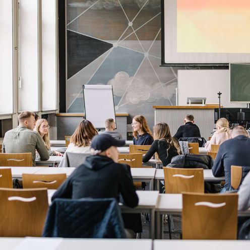 Studierende im Seminar Perspektive aus der hinteren Reihe eines Seminarraums: Studierende sitzen in an mehren Tischreihen verteilt. Einige unterhalten sich miteinander. Vorne im Raum ist eine Leinwand zu sehen, auf die eine Präsentation projiziert ist.