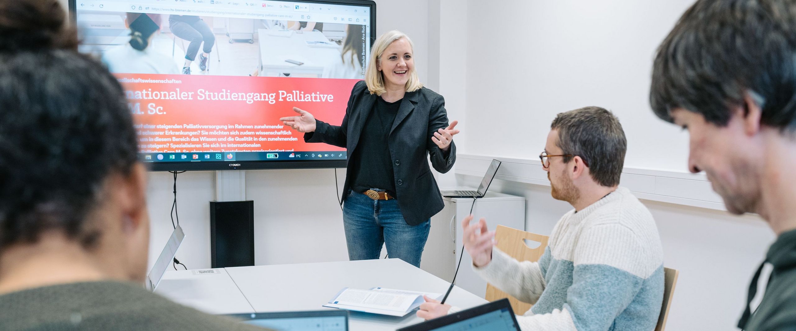 Auf dem Bild ist eine Seminarsituation in kleiner Lerngruppe angemeldet. Die Professorin steht vor einer digitalen Tafel und eine kleine Gruppe Studierende sitzt an Rechnern und hört zu. 