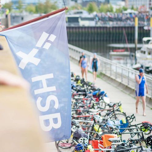 Fahne HSB Gewoba Triathlon Eine blaue Fahne mit dem HSB Logo wird von einem Mann gehalten. Im Hintergrund ist der Marina Hafen in Bremen-Überseestadt zu sehen.