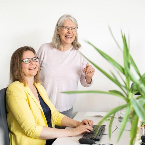 Arbeitsplätze Auf dem Foto sind zwei Frauen zu sehen. Eine sitzt an einem Schreibtisch vor einem Computer, die andere steht hinter dem Stuhl. Rechts im Bild ist eine grüne Palme. Beide Frauen lachen und schauen knapp an der Kamera vorbei.