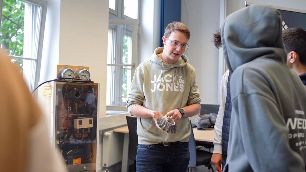 Auf dem Bild ist ein Student der HSB zu sehen. Er hält einen Filter einer Maschine, die neben ihm steht in der Hand. Er erklärt Schülern daran etwas. Der Student trägt einen grünen Kaputzenpullover und eine dunkle Jeans. Er hat kurze bauen Haare und trägt eine Brille. 