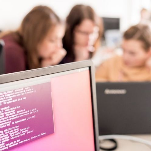 Drei Studentinnen programmieren am Laptop Bildschirm mit Codezeilen im Vordergrund, dahinter unscharf drei Studentinnen, die an einem Laptop sitzen und programmieren.
