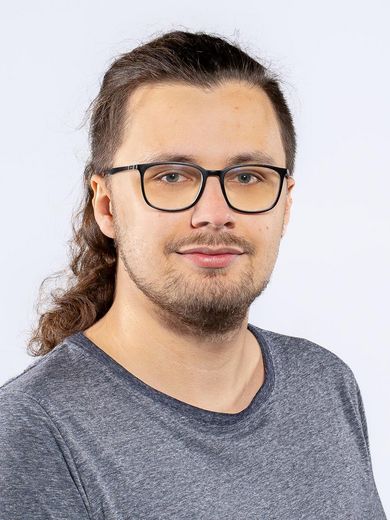 Christopher Lausch Auf dem Bild ist Christopher Lausch zusehen. Er hat lange braune Haare und einen Drei-Tage-Bart. Er trägt eine Brille und ein blaues T-Shirt.