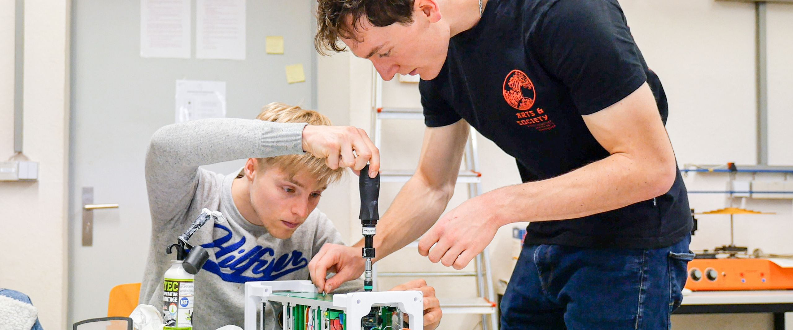 VIBES Belgien_Studierende bei den Vorbereitungen Zwei Studierende schrauben an einem technischen Gerät auf einem Tisch.