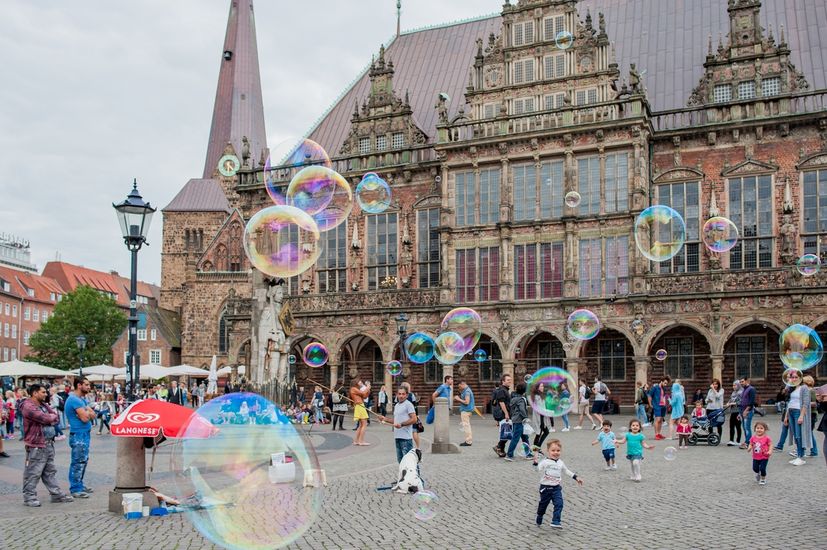 Auf dem Bild ist das Bremer Rathaus zu sehen. Im Vordergrund sind Seifenblasen, spielende Kinder und div. Menschen zu sehen. 