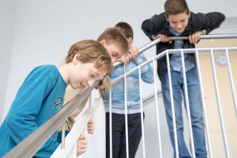 Beobachten Auf dem Bild ist eine Gruppe von Schüler:innen zu sehen, die sich gespannt über das Treppengeländer nach unten beugen, um unten etwas zu beobachten.