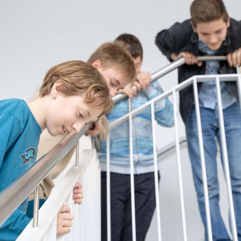 Beobachten Auf dem Bild ist eine Gruppe von Schüler:innen zu sehen, die sich gespannt über das Treppengeländer nach unten beugen, um unten etwas zu beobachten.