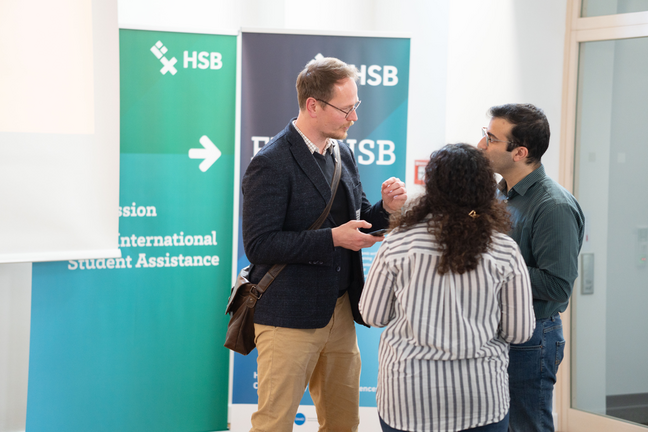 Career Service Drei Menschen stehen sich gegenüber und unterhalten sich. Im Hintergrund hängen HSB Plakate.