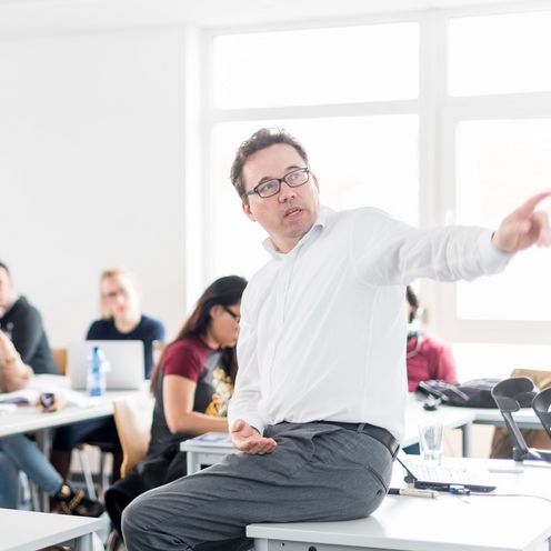 Seminar Professor Giesa Studierende sitzen an Tischen in einem Seminarraum und hören einem Professor zu, der vor ihnen sitzt und etwas erklärt.