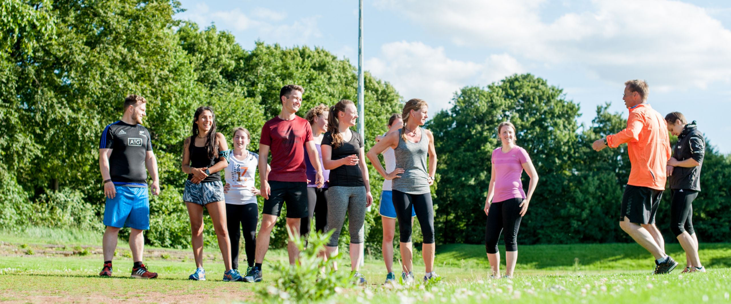 Läufer:innen beim Training Eine Gruppe von Läufer:innen in bunter Sportkleidung steht auf einer Wiese und ist ihrem Trainer zugewandt, der auf der Stelle läuft.