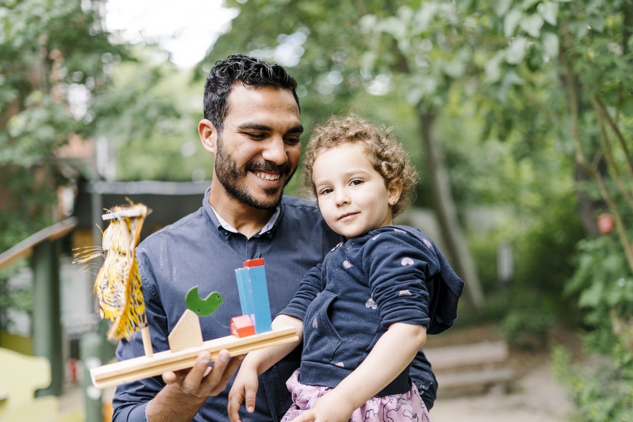 Kind mit Elternteil Ein Kleinkind auf dem Arm seines Elternteils. Das Elternteil hält ein gebasteltes Objekt in der anderen Hand.