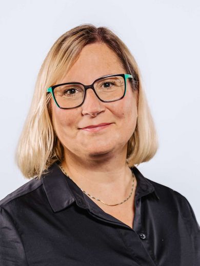 Auf dem Bild ist Ann Pujkiewicz zusehen. Sie hat kinnlanges blondes Haar und trägt eine Brille, sowie eine schwarze Bluse. 