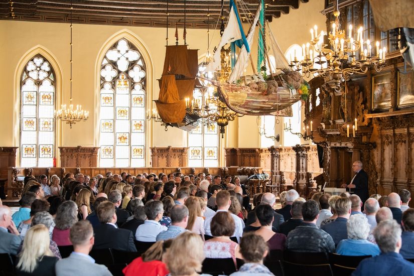 Auf dem Bild ist die obere Halle des Bremer Rathauses zu sehen. Über den Köpfen des sitzenden Publikums hängen große Modelle von Schiffen. Im Hintergrund sind helle Rundbogenfenster mit künstlerischen Verzierungen zu sehen. 