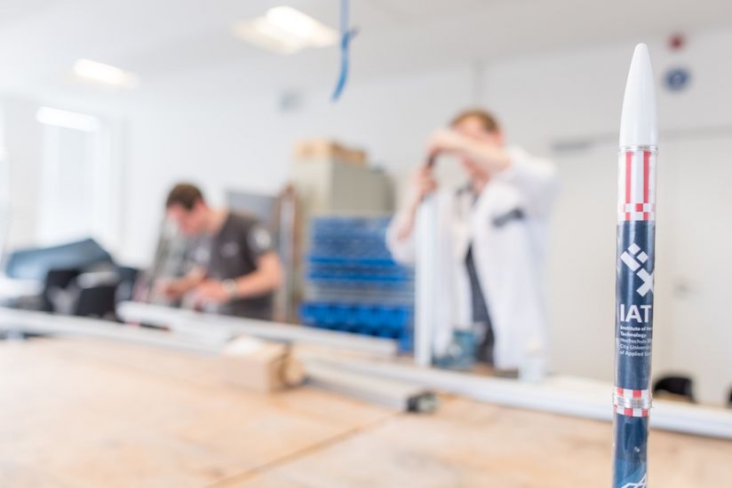 Werkstatt Luft- und Raumfahrt Studierende arbeiten an mechanischen Bauteilen in einer Werkstatt. Im Vordergrund steht das Miniaturmodell einer Rakete.