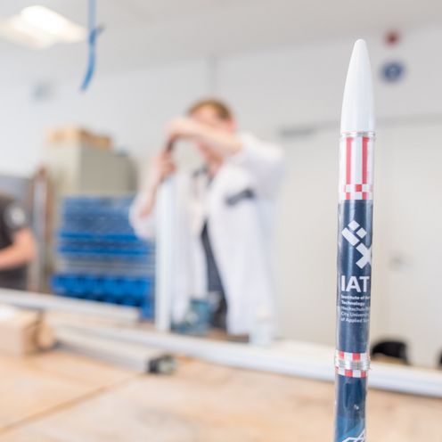 Werkstatt Luft- und Raumfahrt Studierende arbeiten an mechanischen Bauteilen in einer Werkstatt. Im Vordergrund steht das Miniaturmodell einer Rakete.