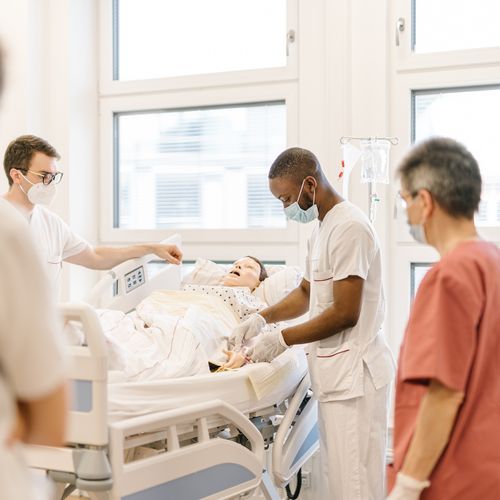 2. Förderphase TCALL Pflege Pflegende stehen an einem Pflegebett, in dem eine Modellpuppe steht.