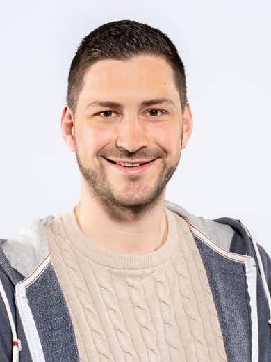 Jonas Unterholzner Auf dem Foto ist Jonas Unterholzner zusehen. Er hat kurze braune Haare und einen Drei-Tage-Bart. Er trägt einen beigen Pullover unter einer blauen Sweatshirtjacke.