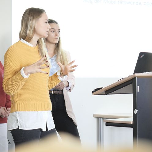 Vortragssituation INA Auf dem Bild sind drei Studentinnen zu sehen, die vor einer Gruppe Menschen einen Vortrag hält. In Hintergrund an einer weißen Wand lässt sich eine Präsentation erahnen.