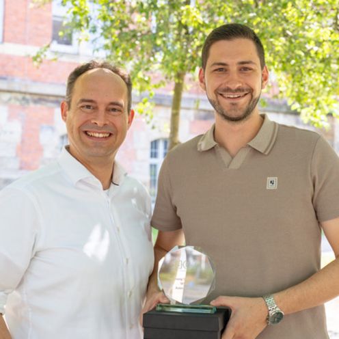 Dirks & Unterholzner mit Young Scientist Award Professor mit Student und dessen Auszeichnung vor Hochschulgebäude