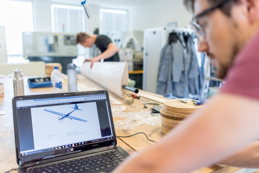 Werkstatt am ZIMT Ein Student betrachtet ein Flugzeugmodell auf einem Laptop, während im Hintergrund ein Student am Bauteil eines Flugzeugmodells arbeitet.